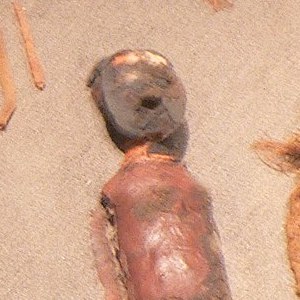The mummified remains of a child lay in a container. The child has a clay mask and red painted clay on their body. 