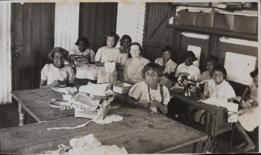 Black and white photo of 10 girls and one teacher seated at tables with needles, fabric, and sewing machines. Girls are dark-skinned. Teacher is white.