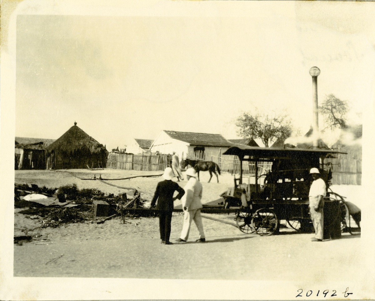 Photo shows three men in pith helmets with a device on a cart in the foreground. A small hut is in the background.