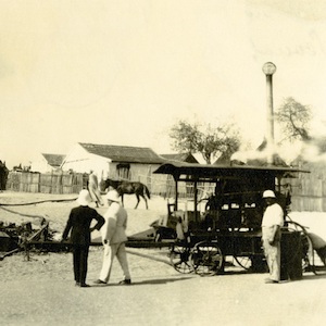 Photo shows three men in pith helmets with a device on a cart in the foreground. A small hut is in the background.