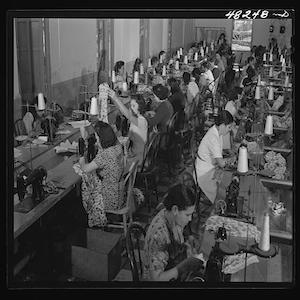 Photo shows women working at sewing machines on both sides of 2 long tables. 