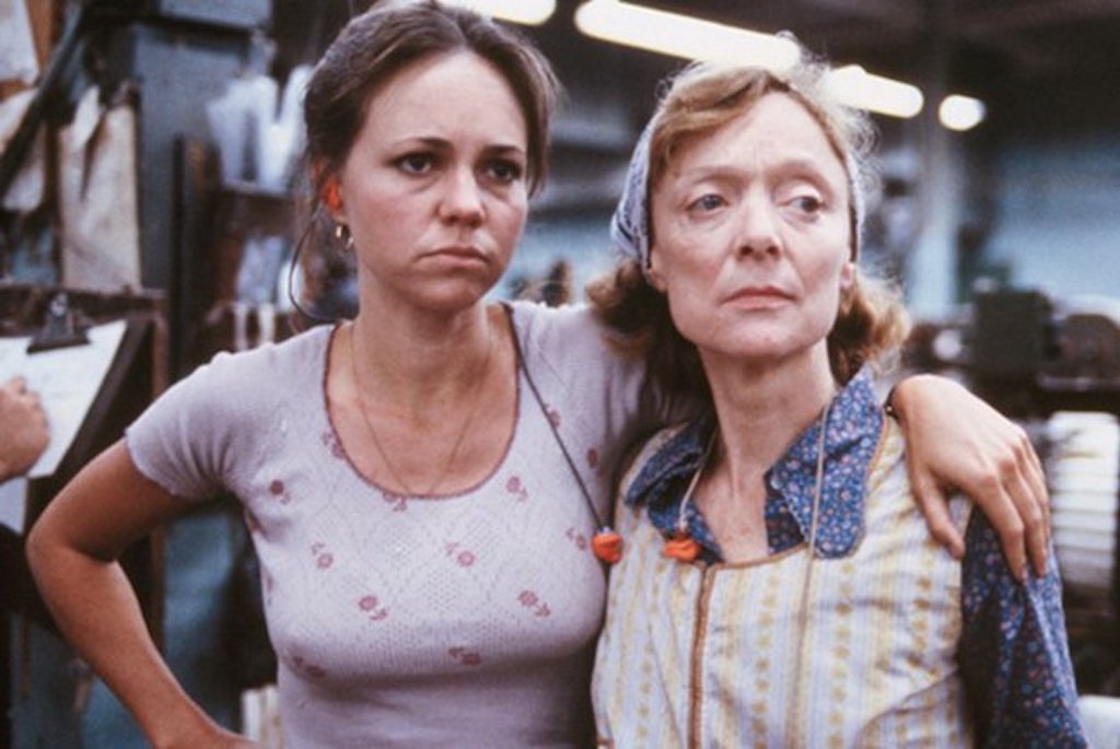 Film still shows two women in a factory. One (portrayed by Sally Field) has her arm around the other.