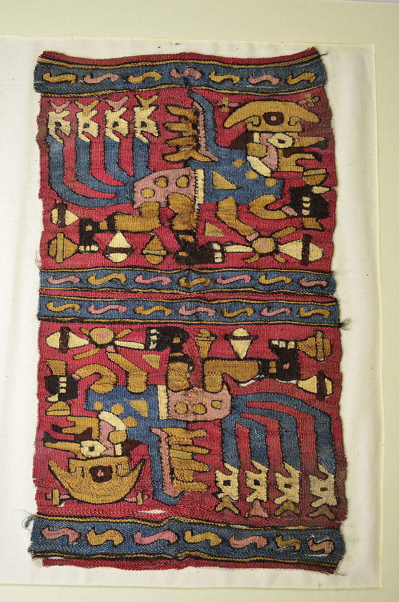 A woven textile with a red background, and blue, yellow, and red geometric patterning. A figure is shown twice, flipped over, and may represent a human-animal hybrid. He wears red and blue clothing, dark colored sandals, and has a golden-colored headpiece. 