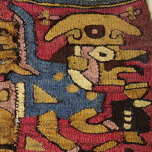 A woven textile with A closeup of a woven textile featuring a figure that may be a human-animal hybrid. He wears red and blue clothing, dark colored sandals, and has a golden-colored headpiece. 