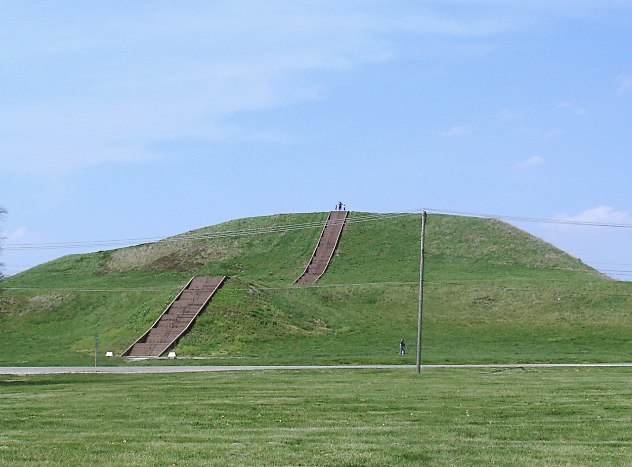 Monks Mound. A large, earthen mound covered in grass set against a blue sky. The mound has stairs with people using them. 