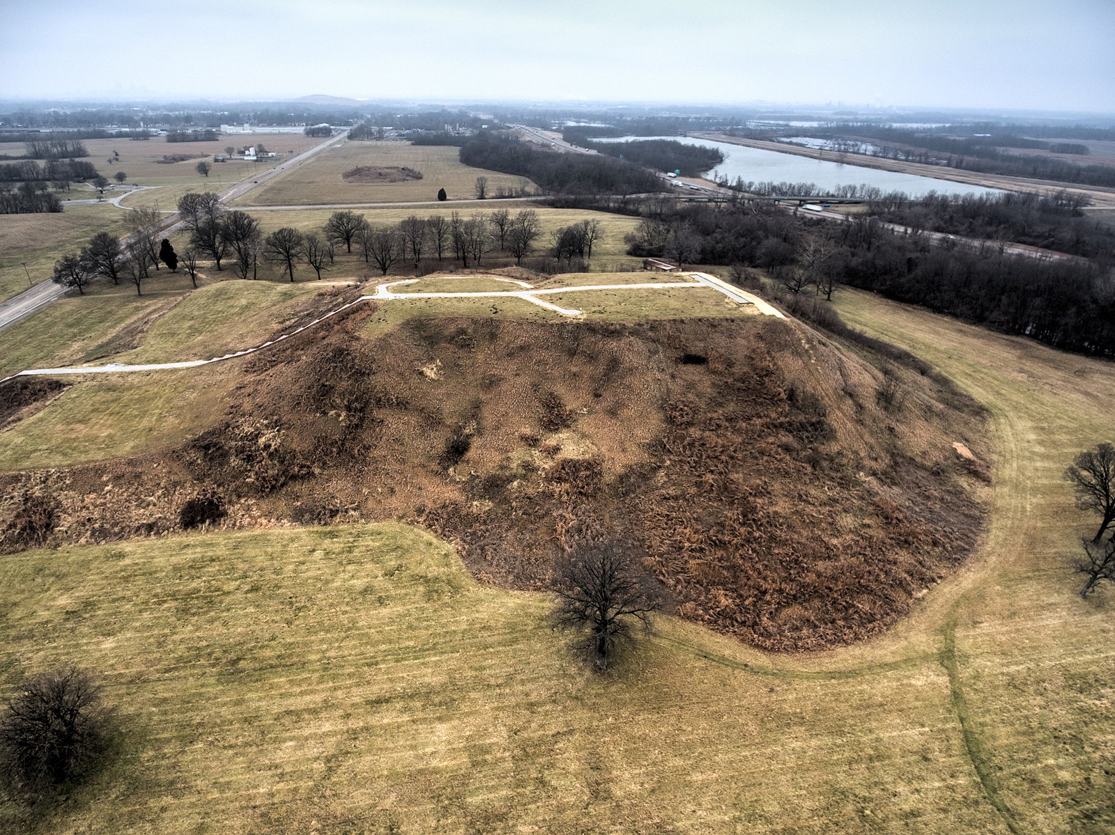 An aerial view of Monks Mound, the largest mound at Cahokia. The brown, earthen mound is in the center and there are roads and waterways in the background. 