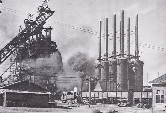 Black and white photo of a factory. Several smoke stacks and smoke is visible.