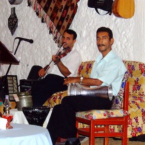 Two men sitting, one has a stringed instrument and one has a drum.