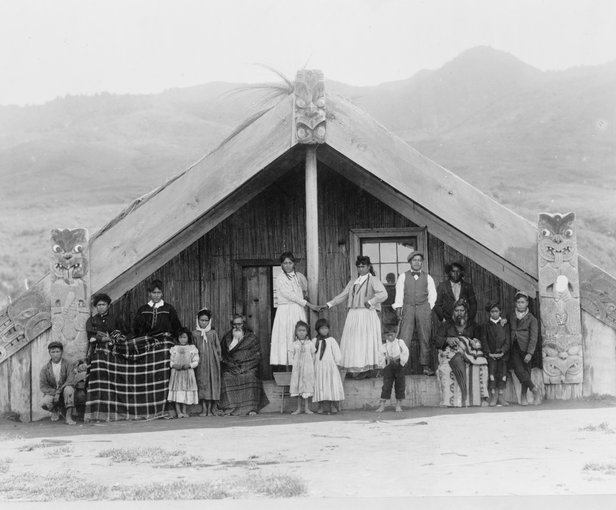 image of a Maori men, women, and children arranged for a group portrait on the porch of a whare or wharenui (meeting house) in New Zealand