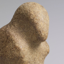 A tan-colored stone figure loosely shaped like a bird with a head and beak and two protruding wings on the side. The figure has a flat bottom. 