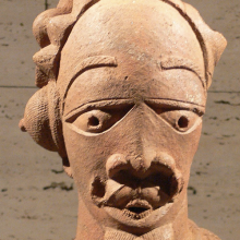 A terracotta sculpture of a male figure with defined facial features, a hairstyle with multiple buns and caps over the ears, and numerous necklaces and other jewelry.