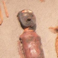 The mummified remains of a child lay in a container. The child has a clay mask and red painted clay on their body. 