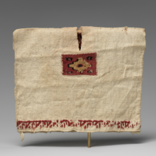 A tan colored tunic stretched to show the height and width. The tunic has a simple slit for the neck, a red and brown geometric design along the chest, and red embroidery along the hem. 