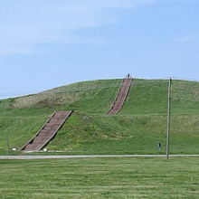 A large, earthen mound covered in grass set against a blue sky. The mound has stairs with people using them. 