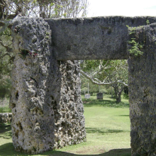 A stone monument with two stones acting as posts and a third stone sitting vertically atop the others. The monument sits on grass and there are trees seen in the background. 